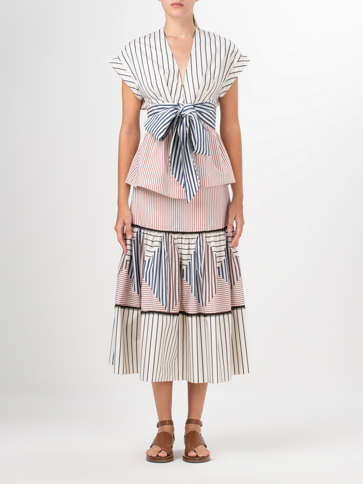 A person models the Elia Blouse by Silvia Tcherassi US, a sleeveless cotton dress featuring vertical stripes in white, black, and red with a large waist bow and layered skirt, paired with brown flat sandals against a plain white background.