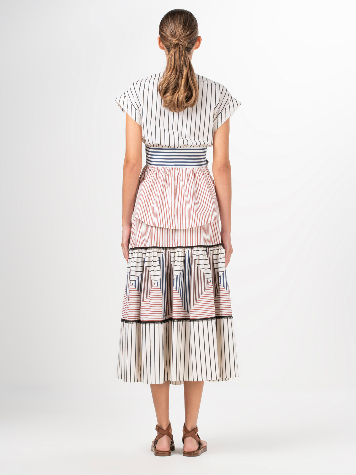 A person with long hair wears the Silvia Tcherassi US Elia Blouse made from cotton fabric with black, white, and pink vertical stripes. The pleated skirt section offers contrasting colors, paired with brown strappy sandals.
