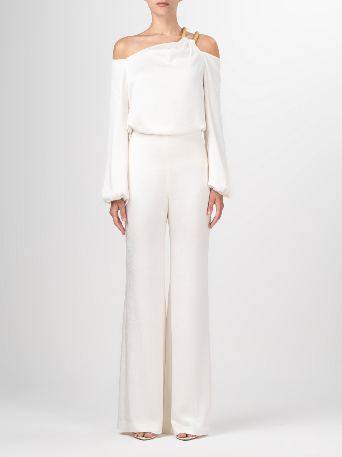 A person is wearing a Barb Blouse by Silvia Tcherassi US; it's an off-the-shoulder white jumpsuit with wide-leg pants and a single long sleeve featuring an asymmetric strap and a gold buckle detail on the shoulder. The person stands against a plain background.